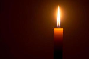 A single burning candle flame or light glowing on a yellow candle isolated on dark red or brown background on table in church for Christmas, funeral and memorial service photo