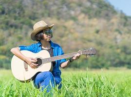 Women short hair wear hat and sunglasses sit playing guitar in grass field photo