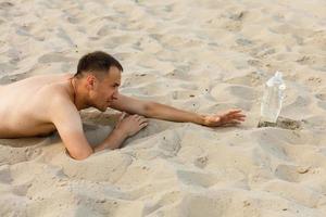 hombre que sufre de sed perdido en el desierto, arrastrándose por la arena hasta la botella con agua en un día caluroso y soleado - supervivencia en la naturaleza. foto