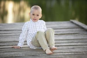 un niño guapo se sienta en un puente de madera junto al lago y mira la cámara. foto
