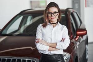 de pie con los brazos cruzados. mujer bonita con falda negra posando cerca de un auto nuevo en el salón del automóvil foto
