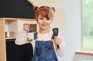 Holding tools in hands and shows it. Little girl in casual clothes have fun by playing with toys on the kitchen photo