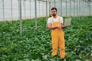 joven trabajador de invernadero con uniforme amarillo con laptop en las manos tiene trabajo dentro del invernadero foto