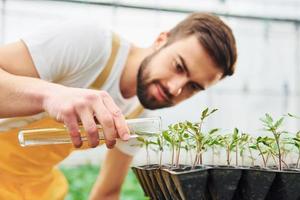 Using test tube and watering plants. Young greenhouse worker in yellow uniform have job inside of hothouse photo