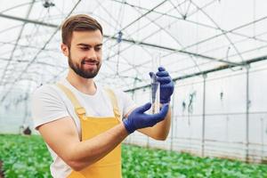 Holds test tube. Young greenhouse worker in yellow uniform have job inside of hothouse photo