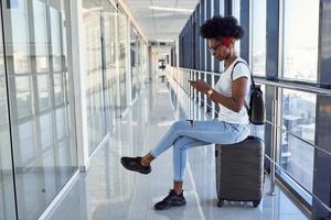 Young african american female passanger in casual clothes is in airport with baggage. Using phone photo