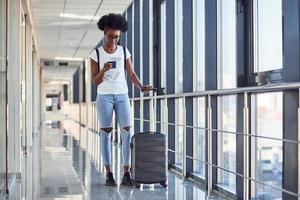 una joven pasajera afroamericana con ropa informal está en el aeropuerto con equipaje. usando el teléfono foto