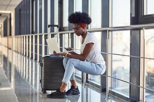 Young african american female passanger in casual clothes is in airport with baggage and using laptop photo
