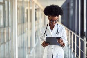 Young african american female doctor in white uniform with stethoscope and notepad standing in corridor photo