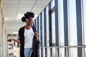 una joven pasajera afroamericana con ropa informal está en el aeropuerto escuchando música con auriculares foto