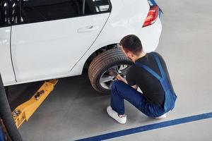 Man in work uniform sitting and changing car wheel indoors. Conception of automobile service photo