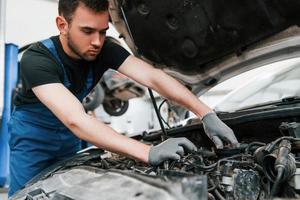 Under the hood. Man in work uniform repairs white automobile indoors. Conception of automobile service photo