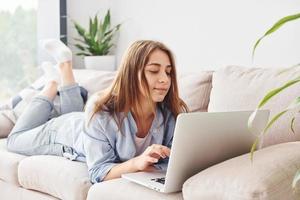 Beautiful young woman in jeans and blue shirt lying down at sofa with laptop photo