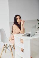 hermosa morena con cabello rizado en ropa de lujo se sienta junto a la mesa con una computadora portátil en casa durante el día foto