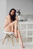 Beautiful brunette with curly hair in luxury clothes sits by table with laptop at home at daytime photo