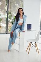 Beautiful brunette with curly hair in blue formal clothes standing indoors near table with laptop on it photo