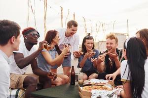 con deliciosa pizza. un grupo de jóvenes con ropa informal tienen una fiesta en la azotea juntos durante el día foto