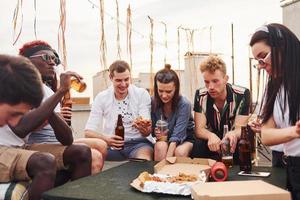 con deliciosa pizza. un grupo de jóvenes con ropa informal tienen una fiesta en la azotea juntos durante el día foto