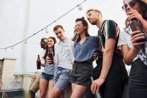 apoyándose en el borde de la azotea con decoraciones. grupo de jóvenes con ropa informal tienen una fiesta juntos durante el día foto