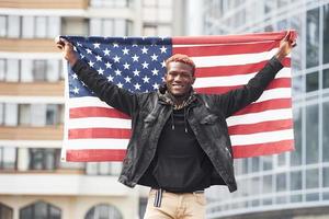 Patriot holding USA flag. Conception of pride and freedom. Young african american man in black jacket outdoors in the city standing against modern business building photo