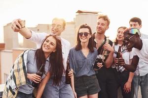 haciendo selfie por teléfono. un grupo de jóvenes con ropa informal tienen una fiesta en la azotea juntos durante el día foto