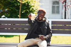 With phone and cup of drink. Young african american man in black jacket outdoors in the city sitting on bench photo