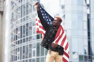 Patriot holding USA flag. Conception of pride and freedom. Young african american man in black jacket outdoors in the city standing against modern business building photo