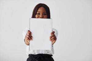 Young african american woman in formal clothes standing with notepad and empty paper in hands indoors photo