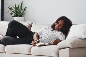 Positive african american woman in white shirt lying down on sofa with notepad in hands photo