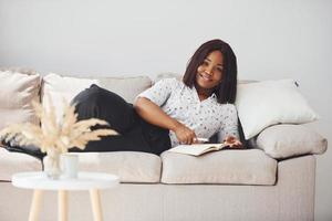 Positive african american woman in white shirt lying down on sofa with notepad in hands photo