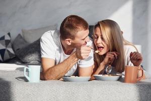una pareja joven y encantadora desayuna en casa mientras se acuesta en la cama foto