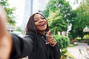 haciendo selfie joven mujer afroamericana con ropa de moda al aire libre en la ciudad cerca de árboles verdes y contra el edificio de negocios foto