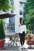 Holding cup of drink in hands. Young afro american woman in white shirt outdoors in the city near green trees and against business building photo