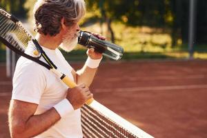 Taking break and drinking water. Senior stylish man in white shirt and black sportive shorts on tennis court photo