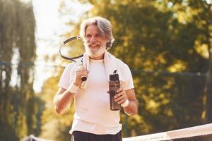 Taking break and drinking water. Senior stylish man in white shirt and black sportive shorts on tennis court photo