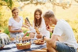 Sitting by picnic table. Group of young people have vacation outdoors in the forest. Conception of weekend and friendship photo