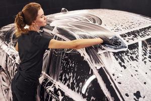 Wipes vehicle that is in white soap. Modern black automobile get cleaned by woman inside of car wash station photo