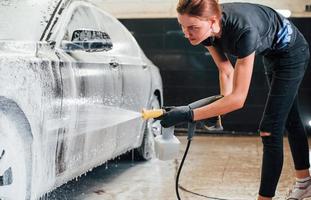 Using high pressure water. Modern black automobile get cleaned by woman inside of car wash station photo
