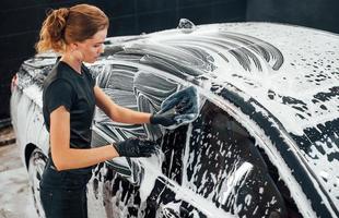 Wipes vehicle that is in white soap. Modern black automobile get cleaned by woman inside of car wash station photo