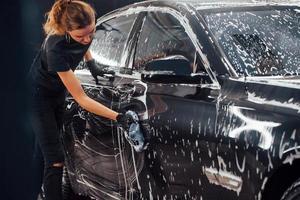 Wipes vehicle that is in white soap. Modern black automobile get cleaned by woman inside of car wash station photo