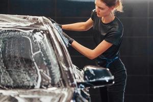 Wipes vehicle that is in white soap. Modern black automobile get cleaned by woman inside of car wash station photo