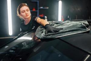 Wipes surface by using towel. Modern black automobile get cleaned by woman inside of car wash station photo