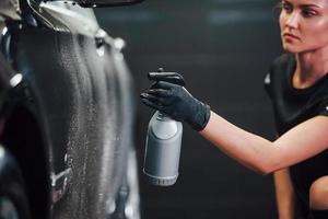 Spraying the vehicle. Modern black automobile get cleaned by woman inside of car wash station photo