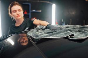 Wipes surface by using towel. Modern black automobile get cleaned by woman inside of car wash station photo
