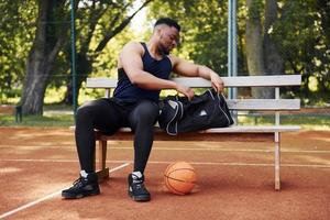 Sits with black bag and preparing for the game. African american man plays basketball on the court outdoors photo