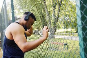 African american guy in wireless headphones leans on the metal mesh of sportive court and takes break photo