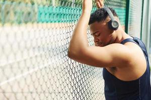 African american guy in wireless headphones leans on the metal mesh of sportive court and takes break photo