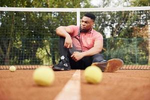 se sienta cerca de la red y toma un descanso. hombre afroamericano con camisa rosa se sienta con raqueta de tenis en la cancha al aire libre foto