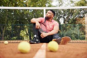 Sits near net and taking a break. African american man in pink shirt sits with tennis racket on the court outdoors photo