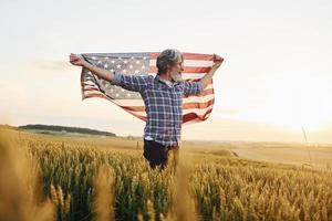 sosteniendo la bandera de estados unidos en las manos. hombre elegante patriótico senior con pelo gris y barba en el campo agrícola foto